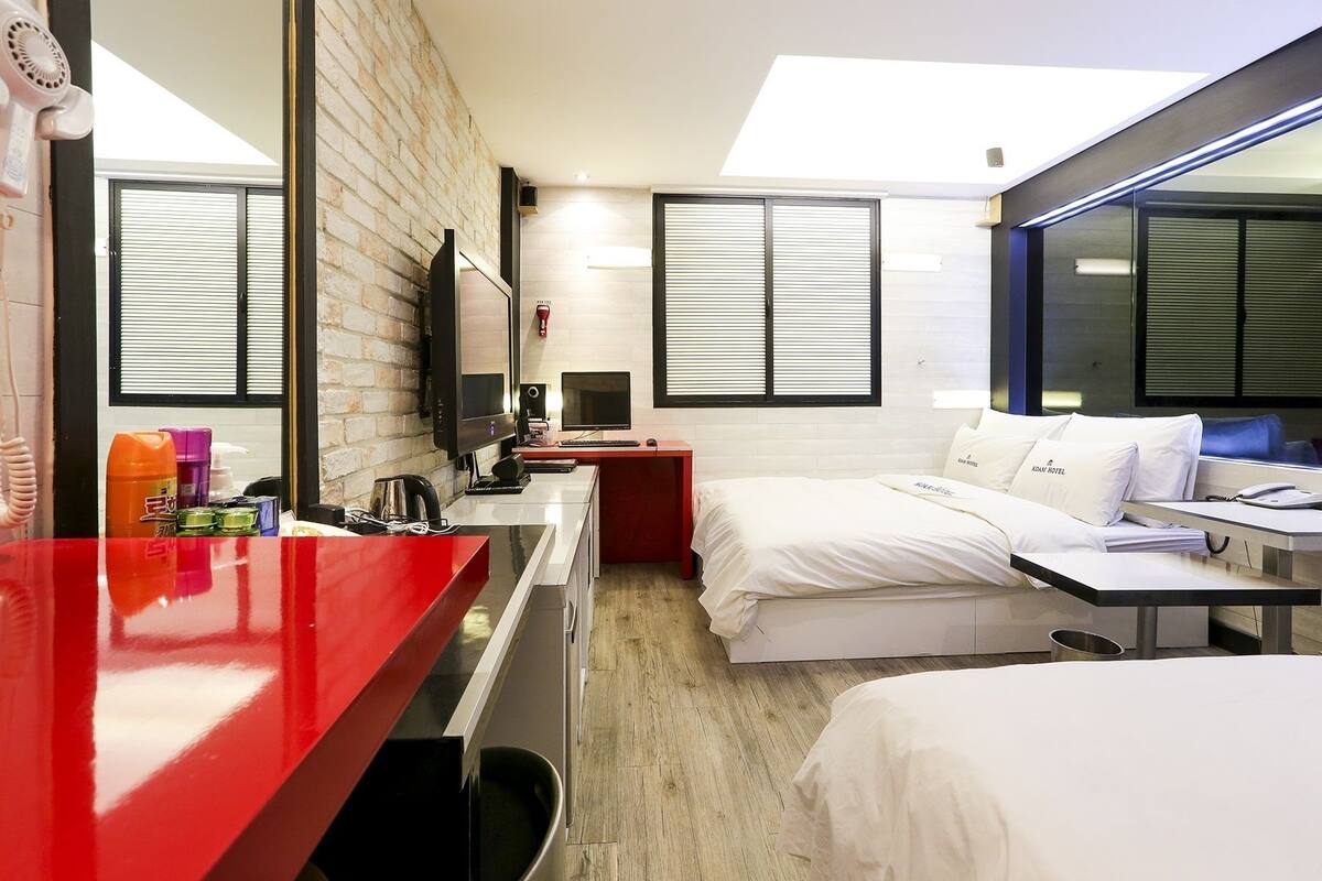 Quarto de hotel com escrivaninha vermelha, cama com roupa de cama branca, televisão e janela com persianas.