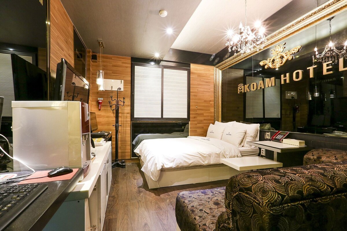 A hotel room with a bed, bedside table, desk with computer, and a large mirror.