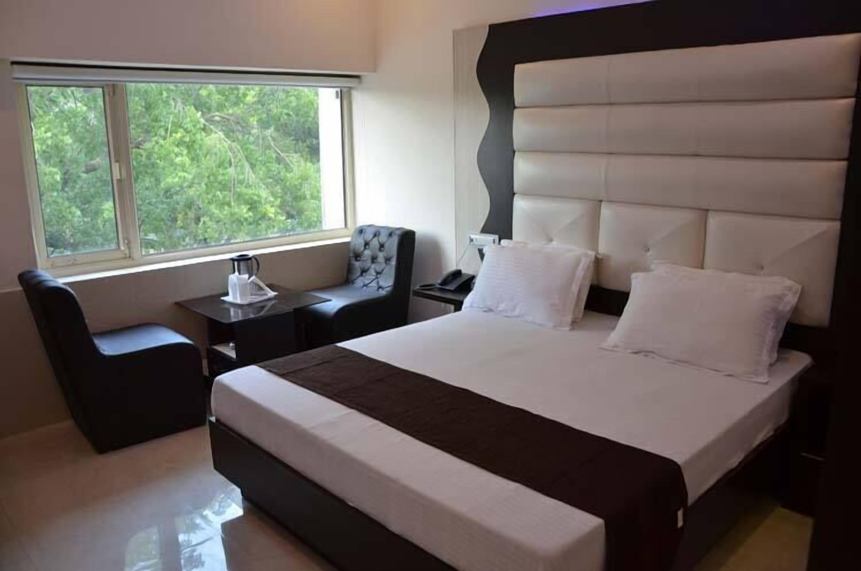 Habitación de hotel moderna con una cama grande, una mesita de noche, una silla y una ventana con vistas al verdor.