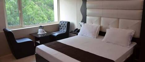 Habitación de hotel moderna con una cama grande, una mesita de noche, una silla y una ventana con vistas al verdor.