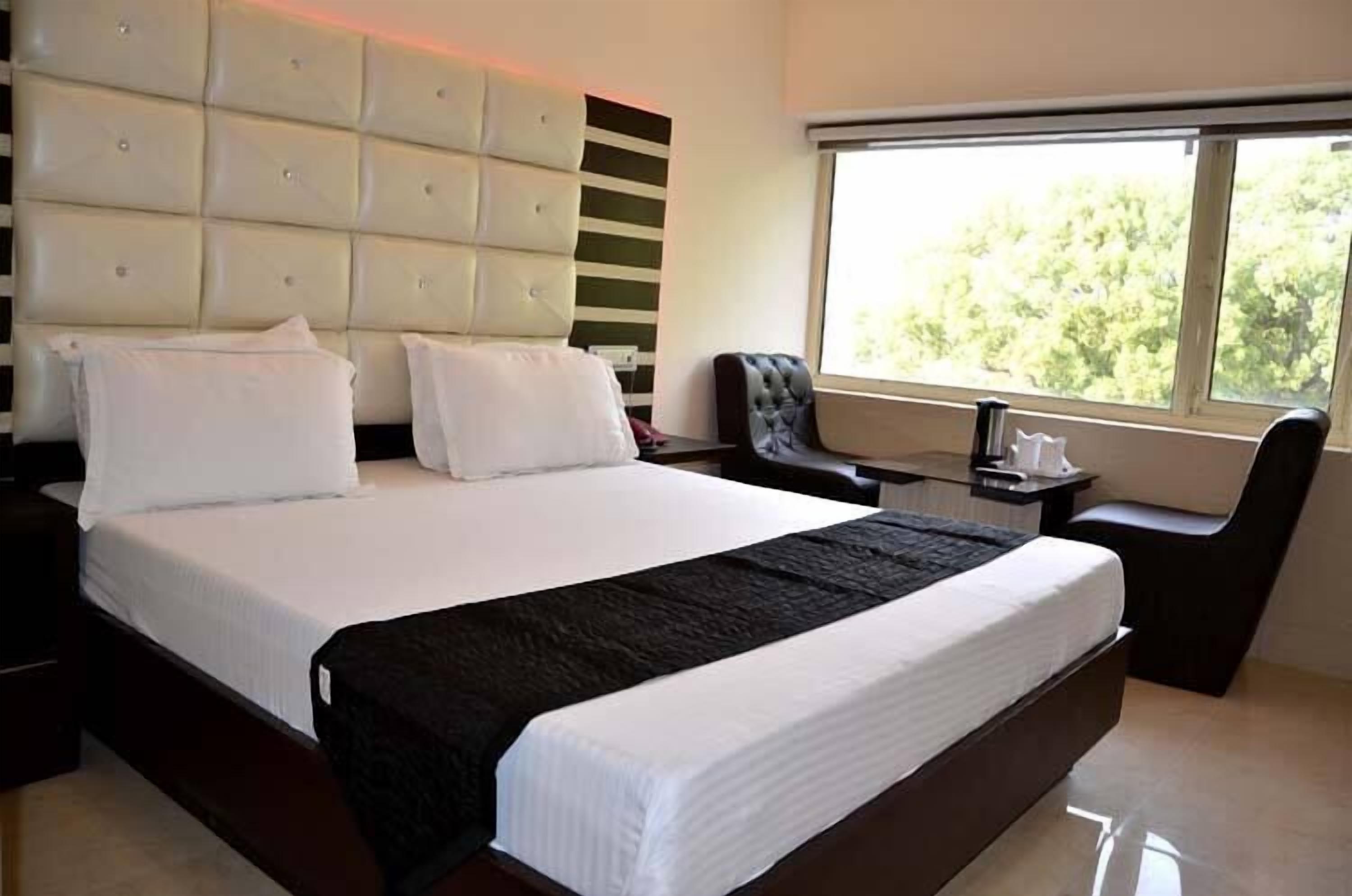 A modern hotel room with a large bed, a padded headboard, a desk, and a window with a view of trees.