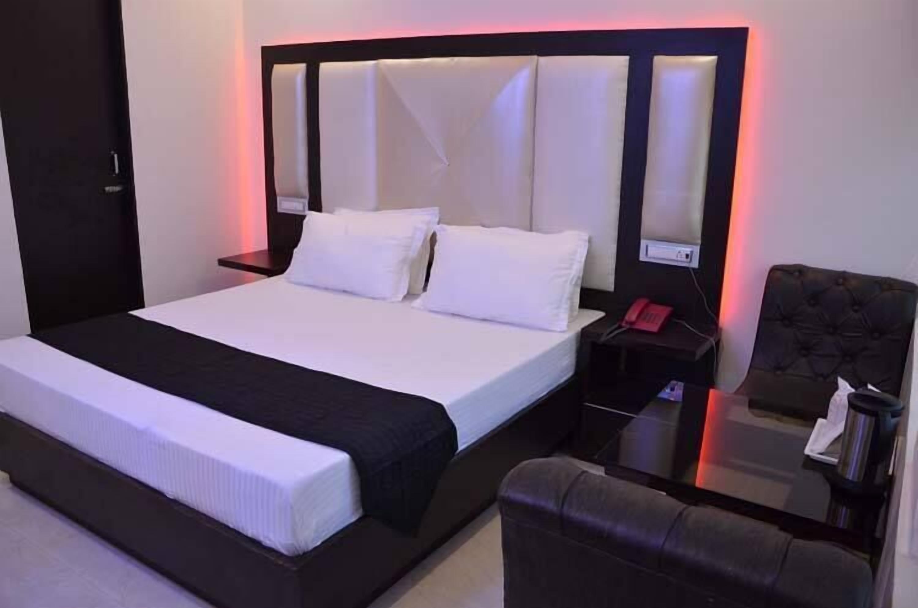 A neatly made bed with white linens and a black headboard. There is a small bedside table with a red phone and a lamp. A dark leather armchair is visible in the corner.