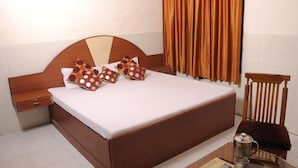 A neatly made bed with a wooden headboard, two pillows with a polka dot pattern, a bedside table, and a chair with a cushion. - Hotel Surya Palace (Chandigarh)