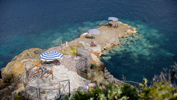 A coastal area with clear blue waters, a rocky platform with two blue and white striped umbrellas, and a few chairs. - Intur Residence (Marciana Marina)