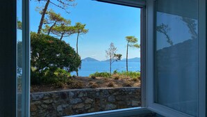A room with a large window overlooking a coastal landscape with trees and a stone wall. - Intur Residence (Marciana Marina)