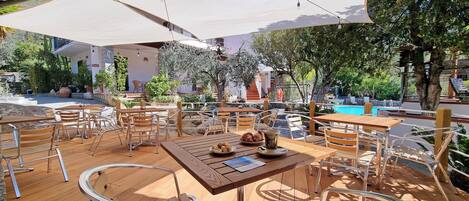 An outdoor seating area with wooden tables and chairs, a pool in the background, and a covered patio.