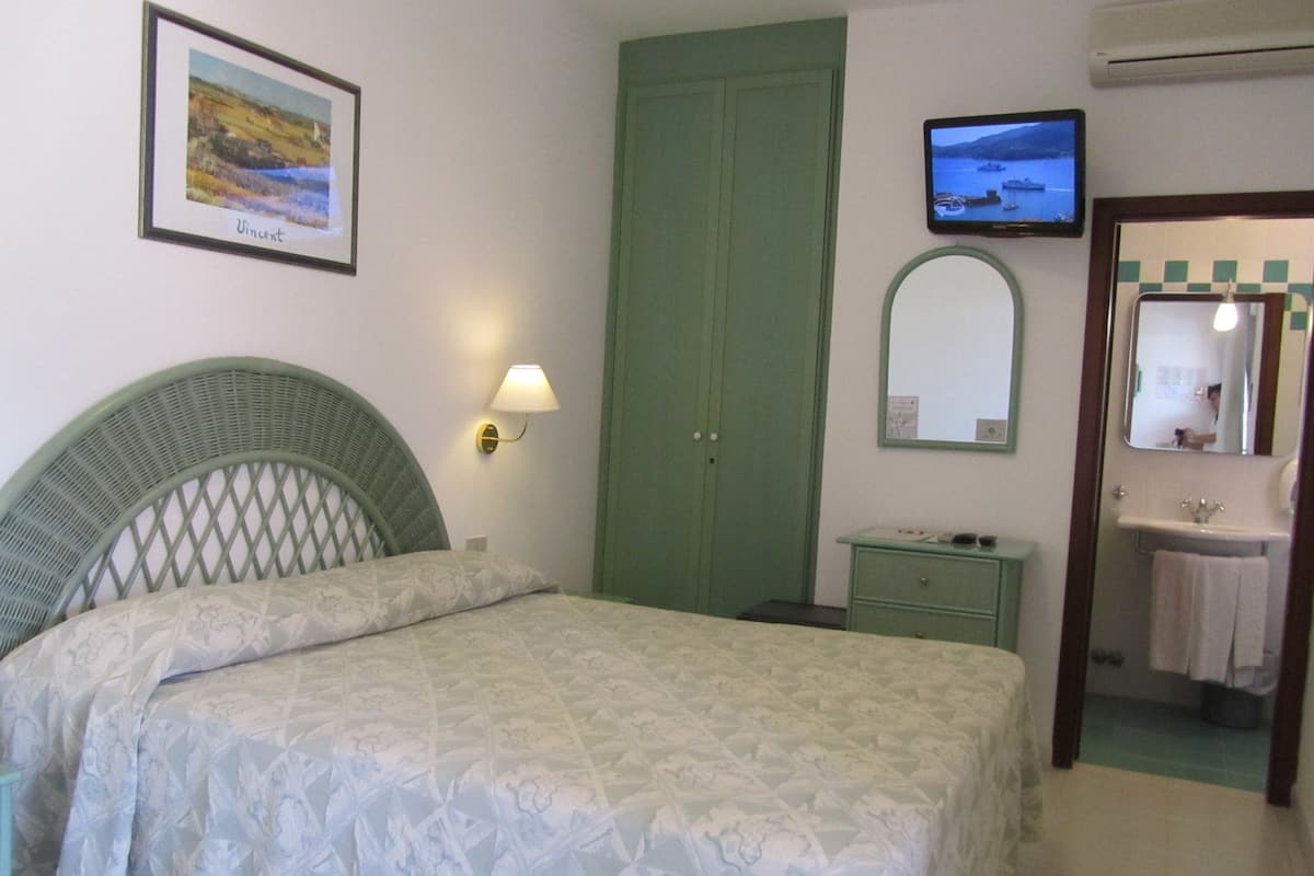 Ein Schlafzimmer mit einem Bett, einem grünen Kleiderschrank, einem kleinen Nachttisch, einem am Wand montierten Fernseher und einem durch eine offene Tür einsehbaren Badezimmer.