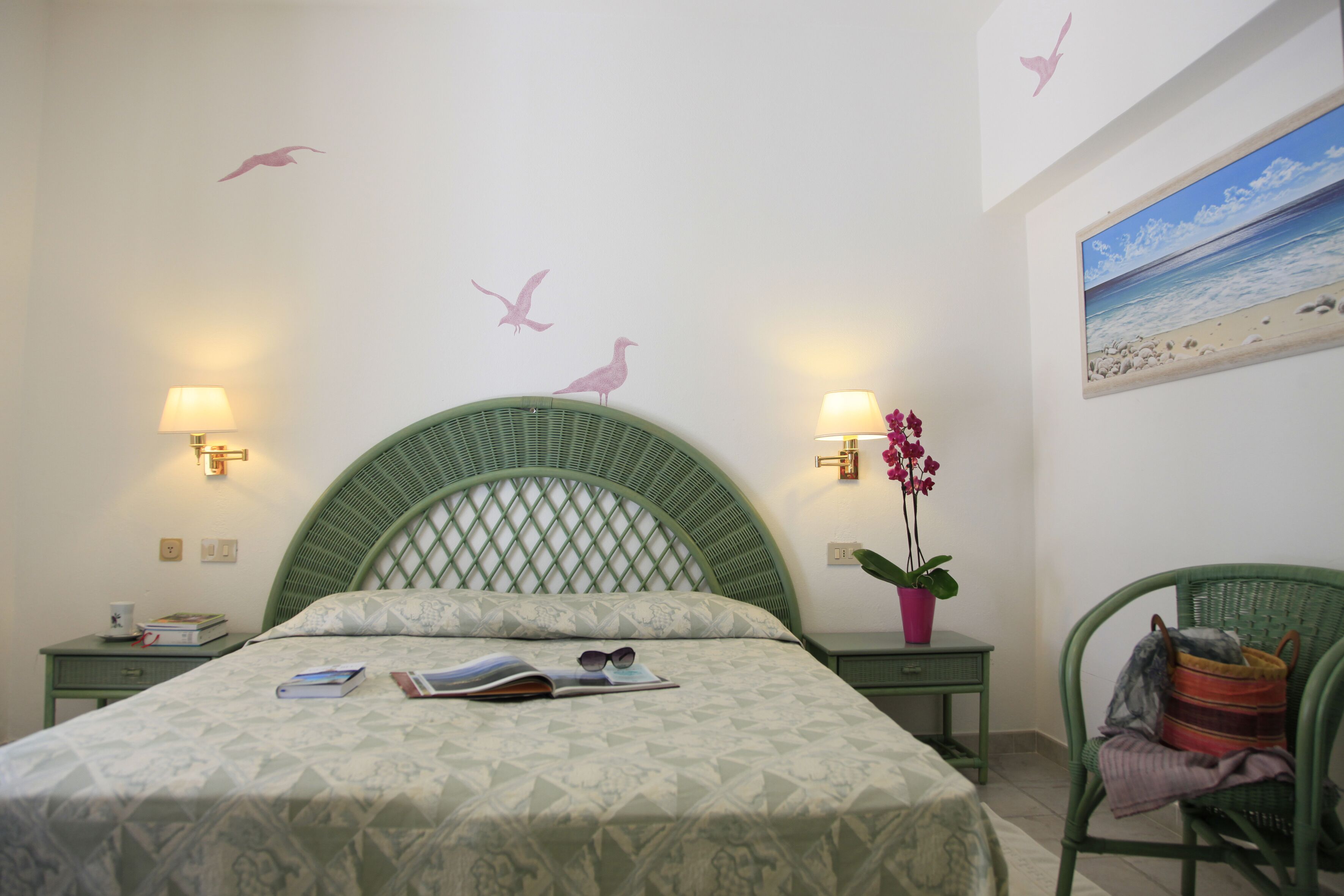 Un dormitorio con cabecera verde, una cama con colcha estampada, dos mesitas de noche con lámparas, una mesita auxiliar con una taza y libros, una silla con una planta, y una pared con un adhesivo de pájaro y un cuadro de una playa.