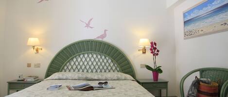 Un dormitorio con cabecera verde, una cama con colcha estampada, dos mesitas de noche con lámparas, una mesita auxiliar con una taza y libros, una silla con una planta, y una pared con un adhesivo de pájaro y un cuadro de una playa.
