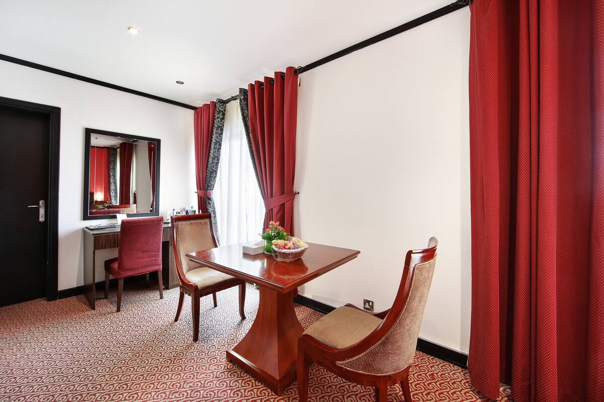 A hotel room with a dining table, two chairs, a mirror, and red curtains.