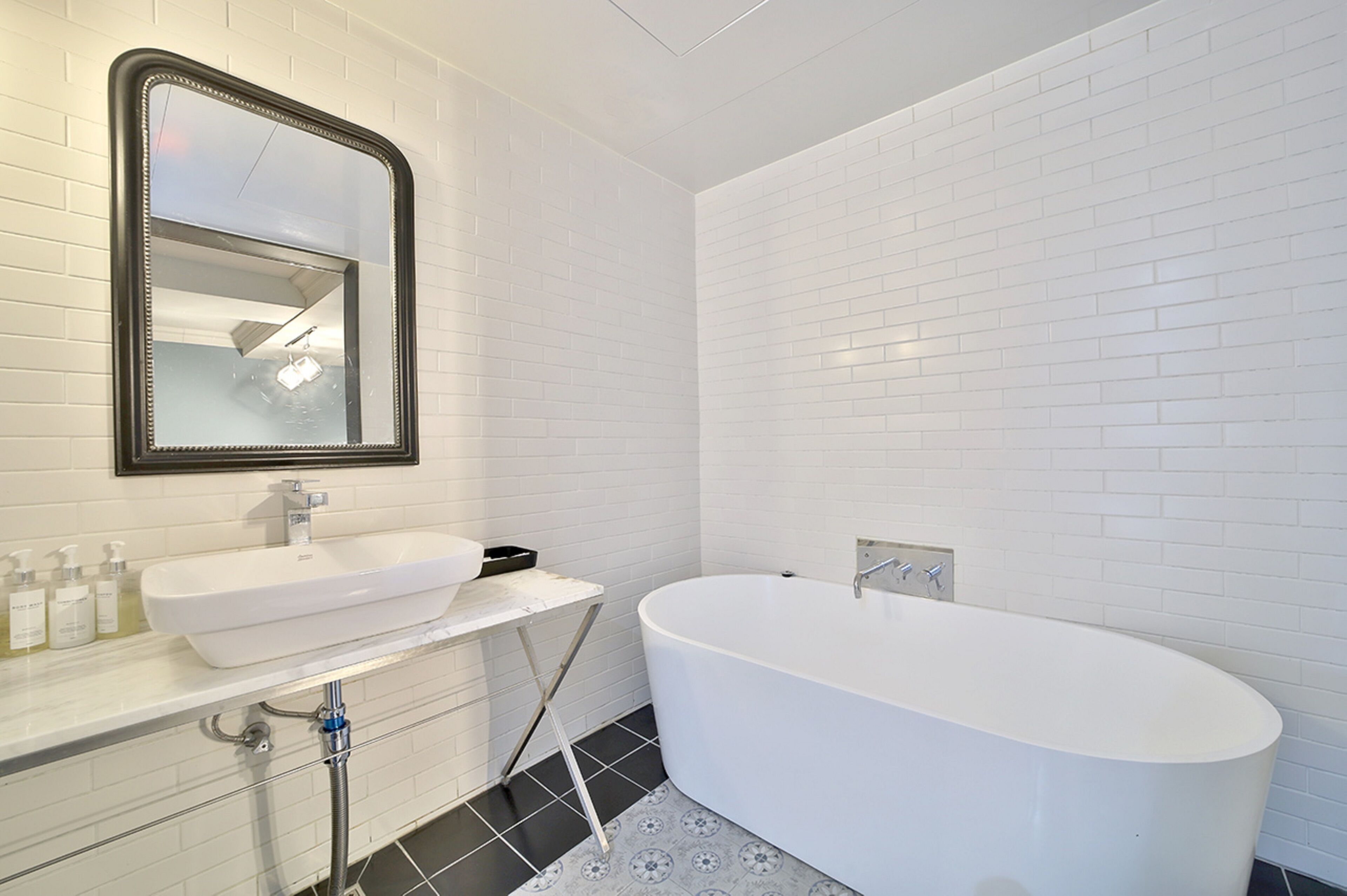 A modern bathroom with white subway tiles, a white freestanding bathtub, a white sink with a mirror above it, and a small shelf with toiletries.