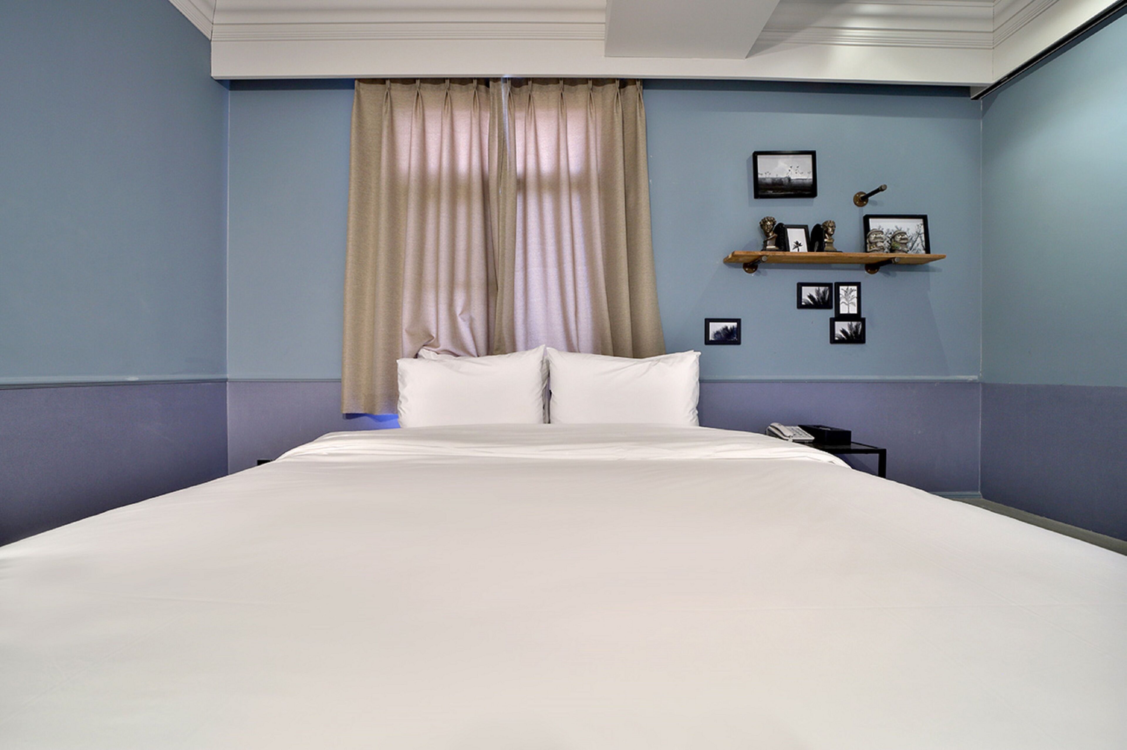 A hotel room with a large bed, beige curtains, a nightstand with a phone, and a wall-mounted shelf with framed pictures.