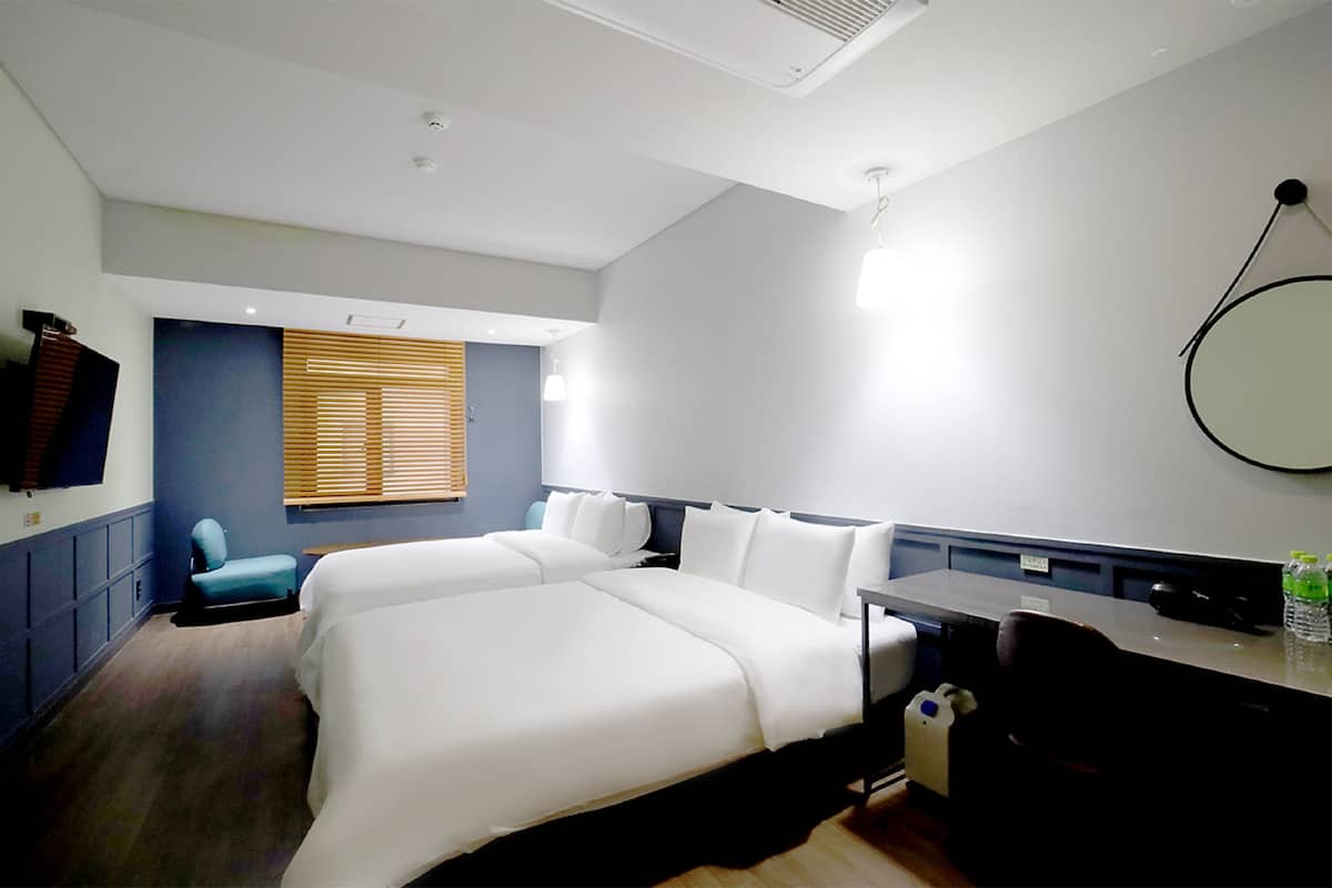 Una habitación de hotel con dos camas, un escritorio, una silla, un televisor y un ventanal grande con persianas.