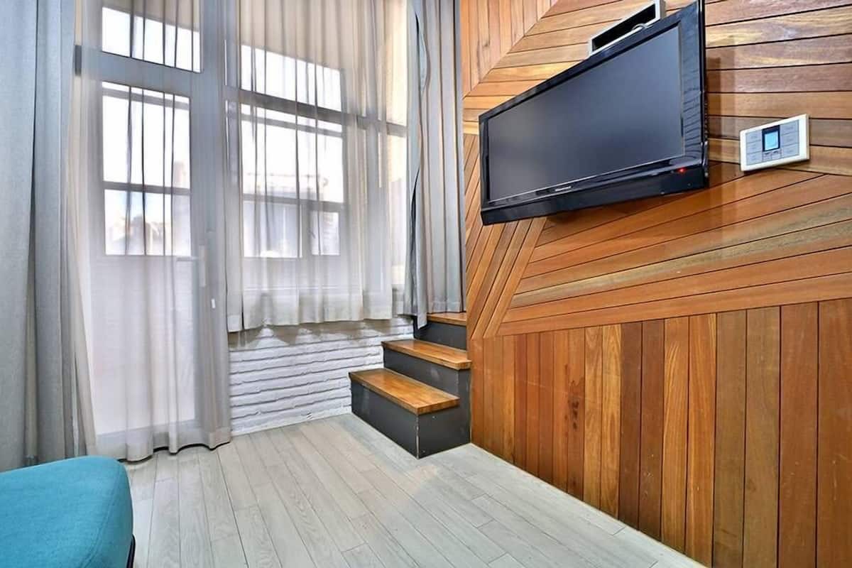 Habitación moderna con televisión montada en una pared de madera, ventana con cortinas translúcidas y sofá azul.
