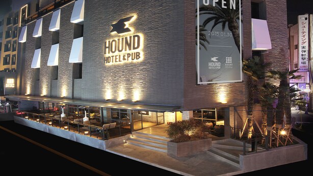 Un hotel con un llamativo letrero que dice 'Gran Inauguración', diseño moderno con techo plano y un área de estar al aire libre visible.
