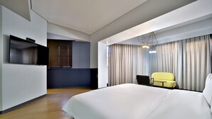 Una habitación de hotel moderna con una cama grande, un televisor de pantalla plana montado en la pared, un pequeño rincón de descanso con un sillón amarillo y un amplio ventanal con cortinas.