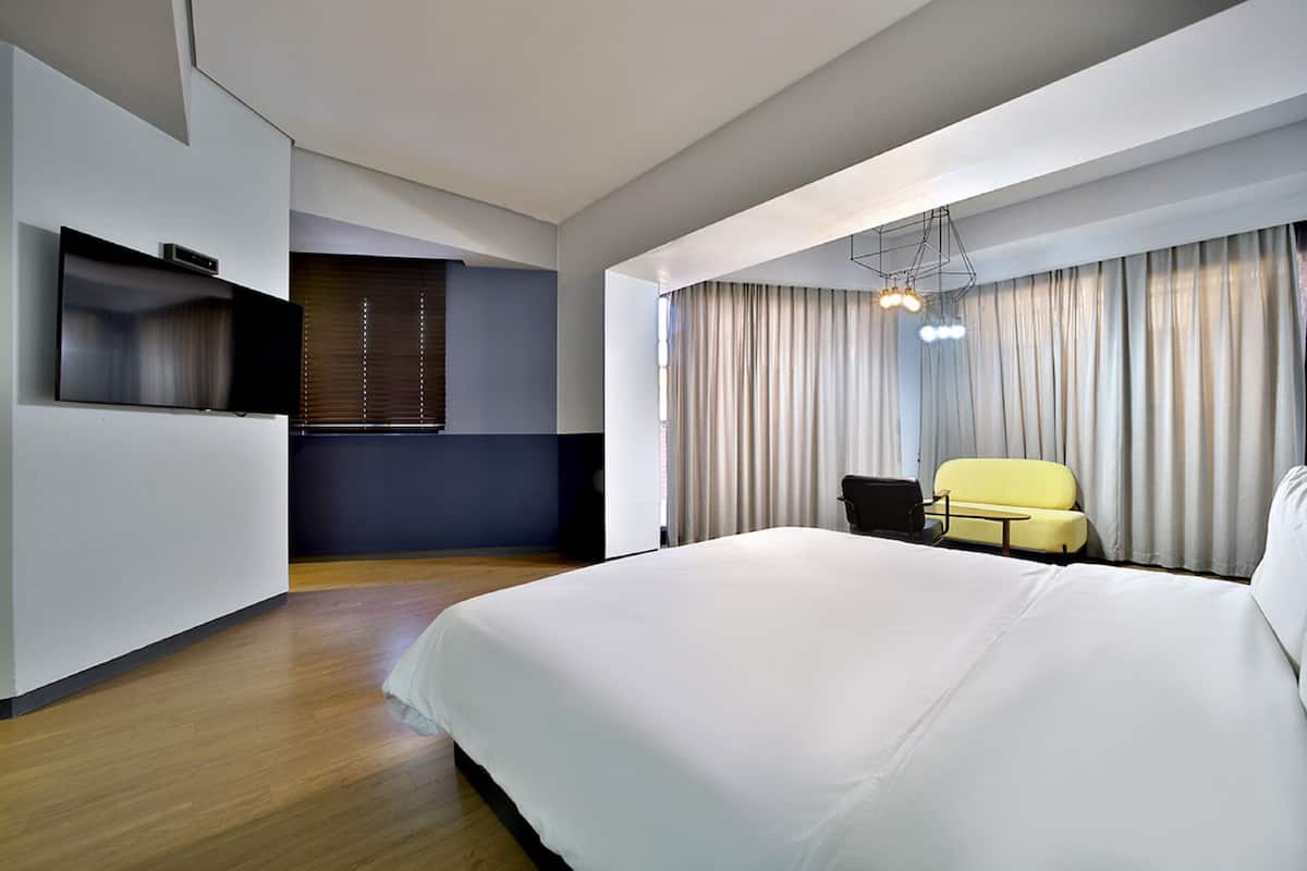 Una habitación de hotel moderna con una cama grande, un televisor de pantalla plana montado en la pared, un pequeño rincón de descanso con un sillón amarillo y un amplio ventanal con cortinas.