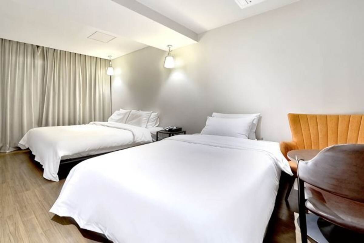 Quarto de hotel com duas camas, uma cadeira, uma mesinha de cabeceira e uma janela grande com cortinas.