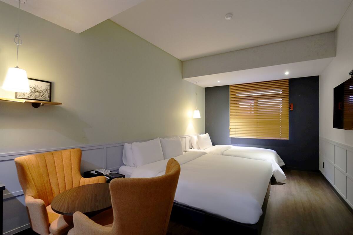 Habitación de hotel con una cama grande, dos sillones, una mesita, televisor de pared y ventana con persianas.