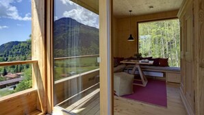 A wooden cabin with a balcony overlooking a mountainous landscape. Inside, there is a seating area with a table and a hanging light fixture.