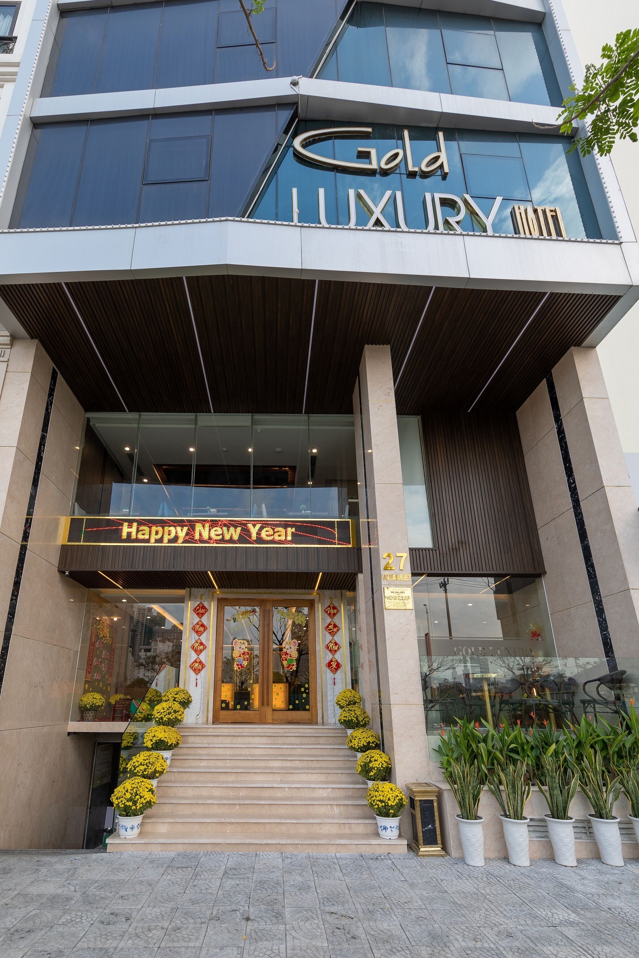 Exterior del Hotel Gold Luxury con un cartel de 'Happy New Year', una escalera con plantas en macetas y un diseño arquitectónico moderno.