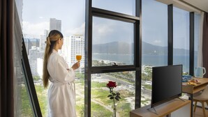 Una mujer con bata blanca, de pie junto a una ventana con un florero de rosas, sosteniendo un vaso de jugo de naranja.