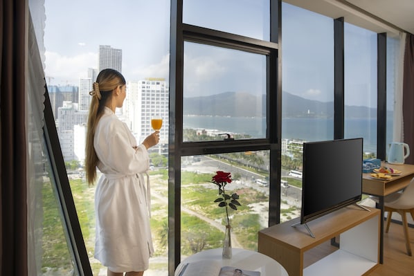 A woman in a white robe standing by a window with a rose vase, holding a glass of orange juice. - Solara Diamond Hotel (Da Nang)
