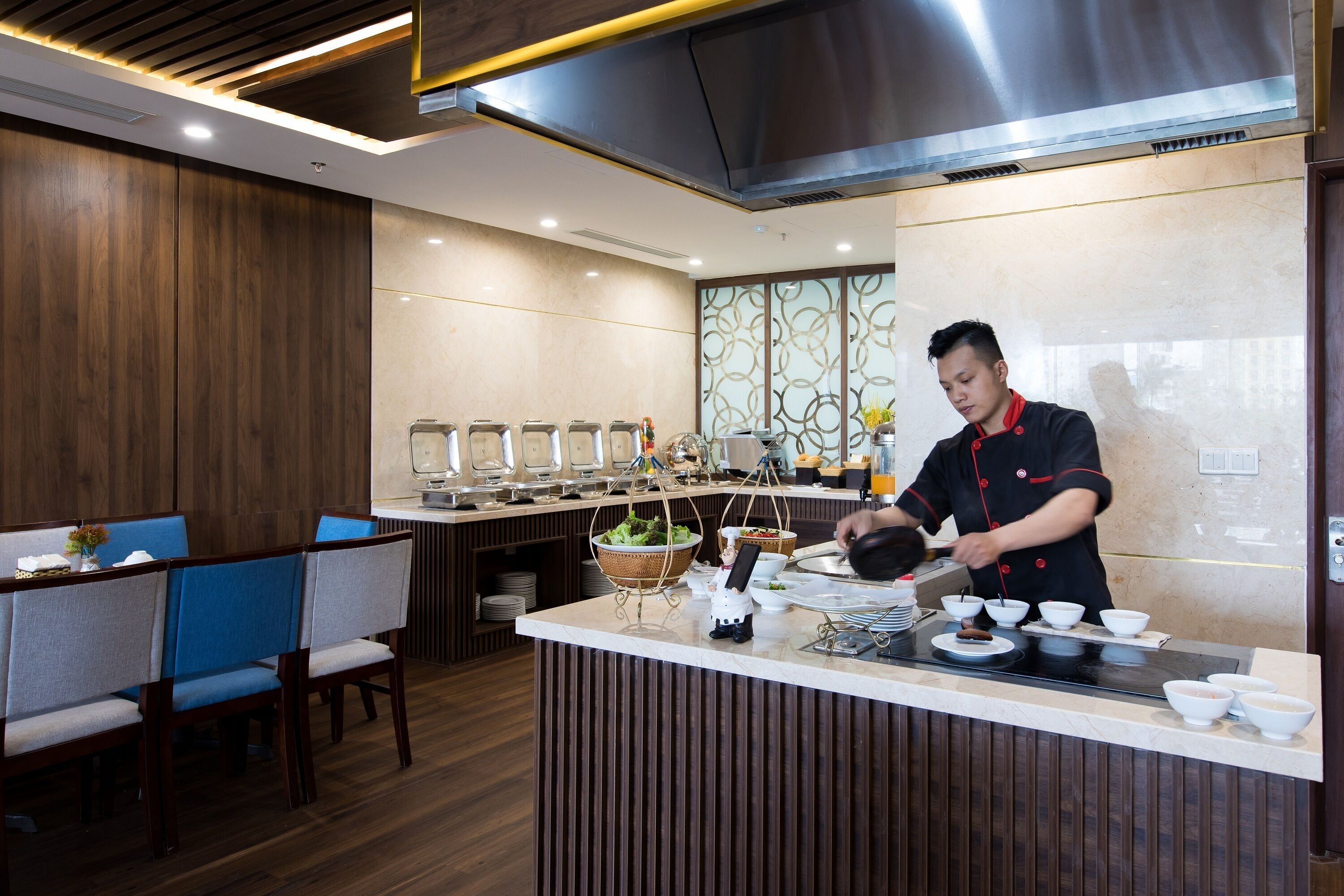 Cocina moderna con un chef preparando comida, un bufé con diversos platos y un área de asientos con sillas azules.