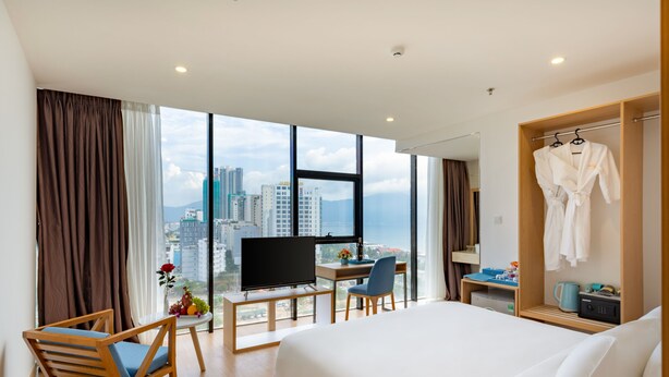 A hotel room with a large bed, a desk with a computer, a chair, a TV, and a view of the city. - Solara Diamond Hotel (Da Nang)