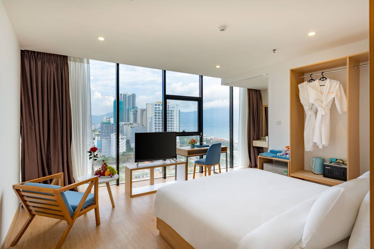 A hotel room with a large bed, a desk with a computer, a chair, a TV, and a view of the city.
