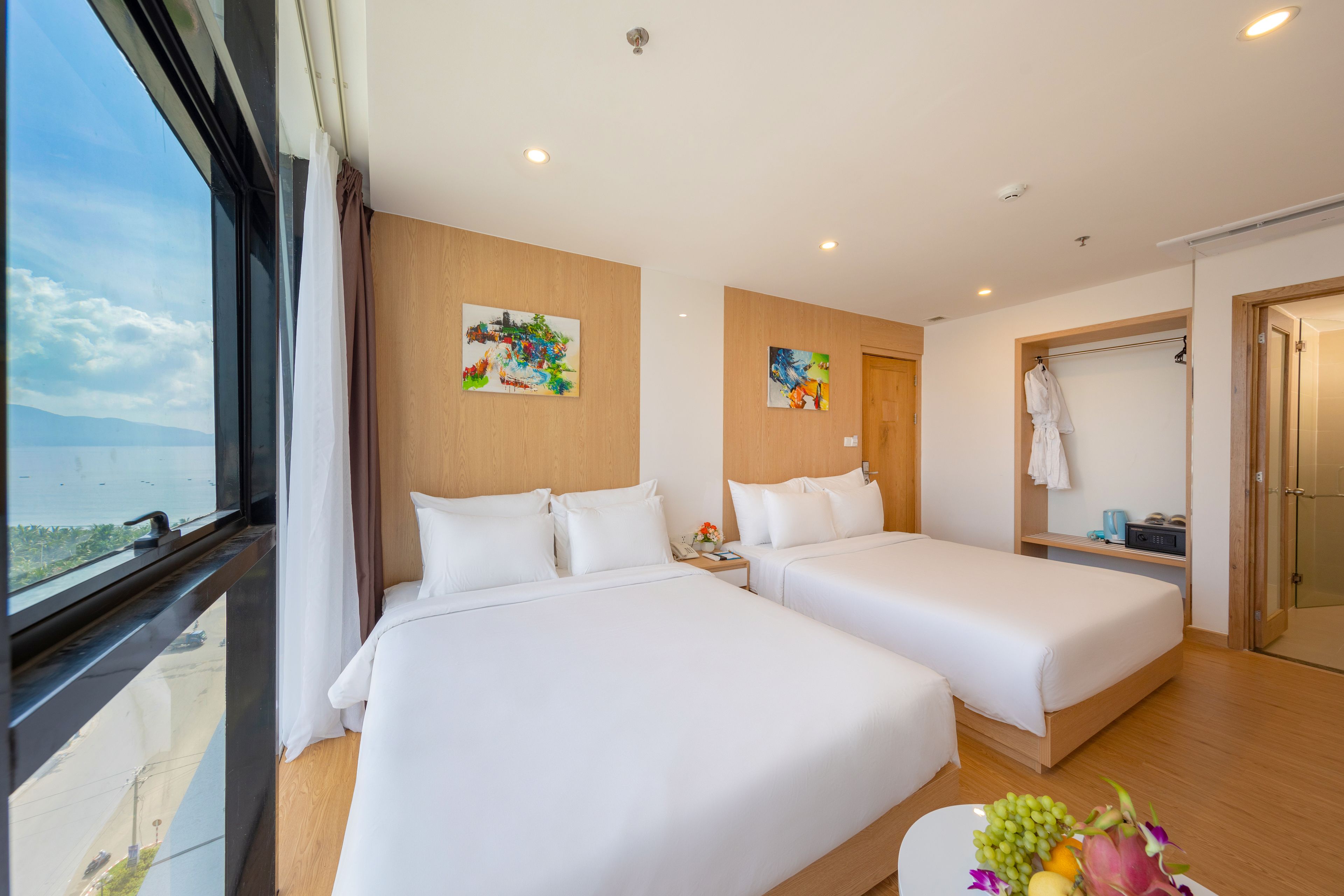 Una habitación de hotel con dos camas, una ventana con vistas, obras de arte en las paredes y un baño con perchero para bata.