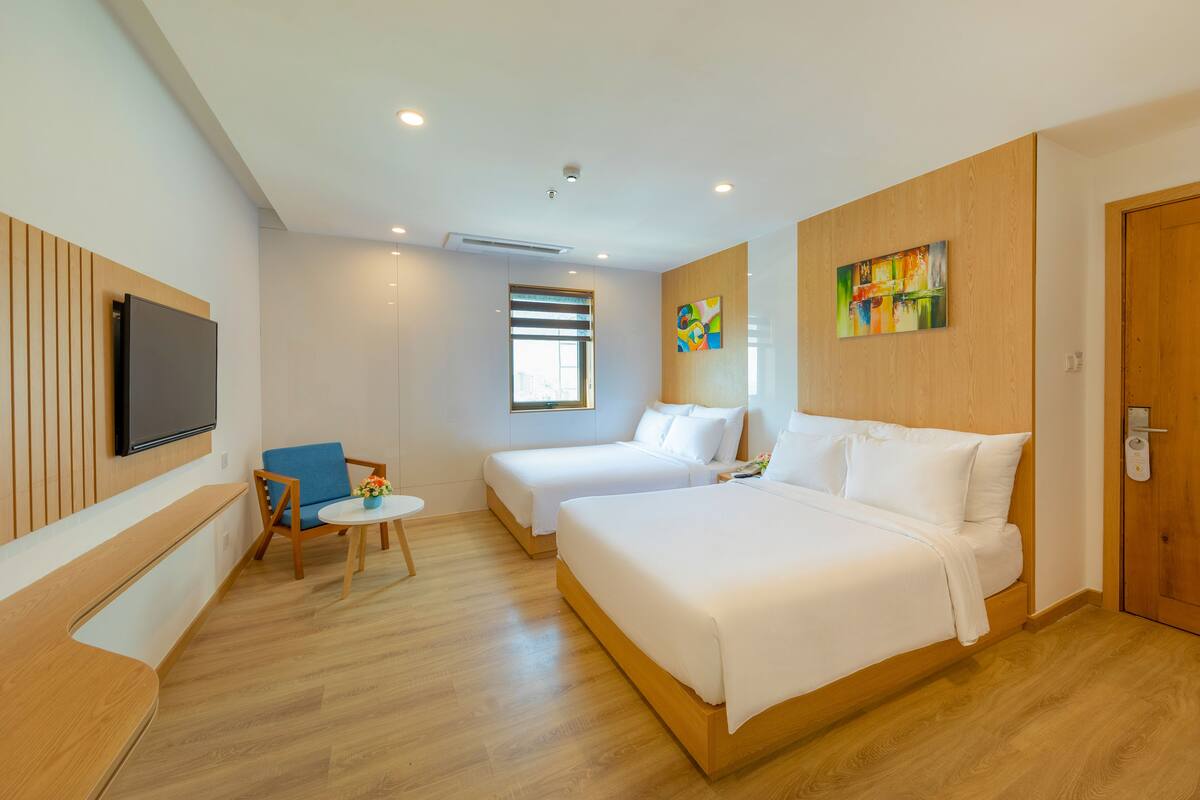 A hotel room with two beds, a TV mounted on the wall, a small table with a chair, and a window with blinds.