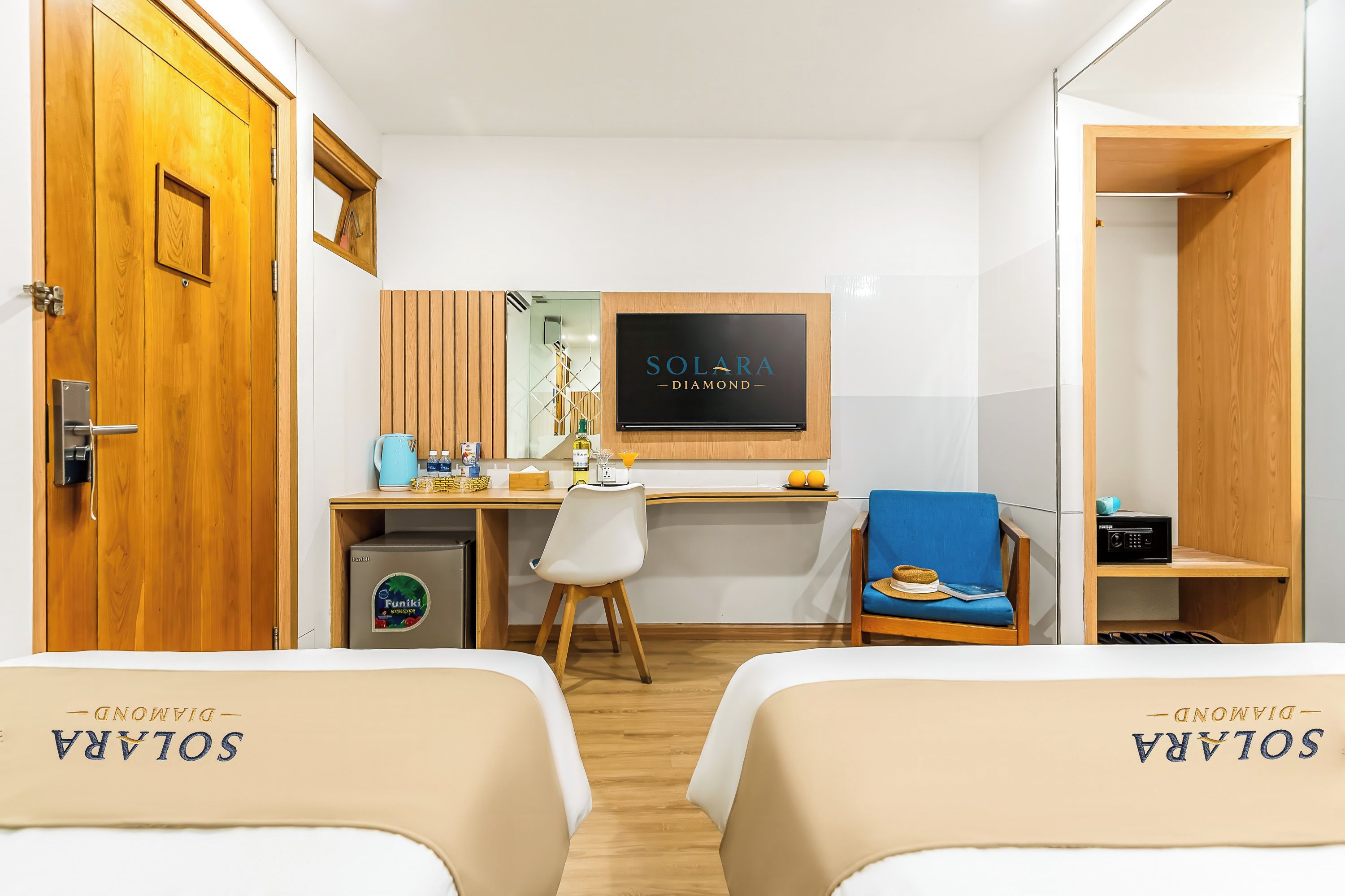 A hotel room with two beds labeled 'SOLARA DIAMOND', a desk with a computer, a chair, a TV, and a minibar.