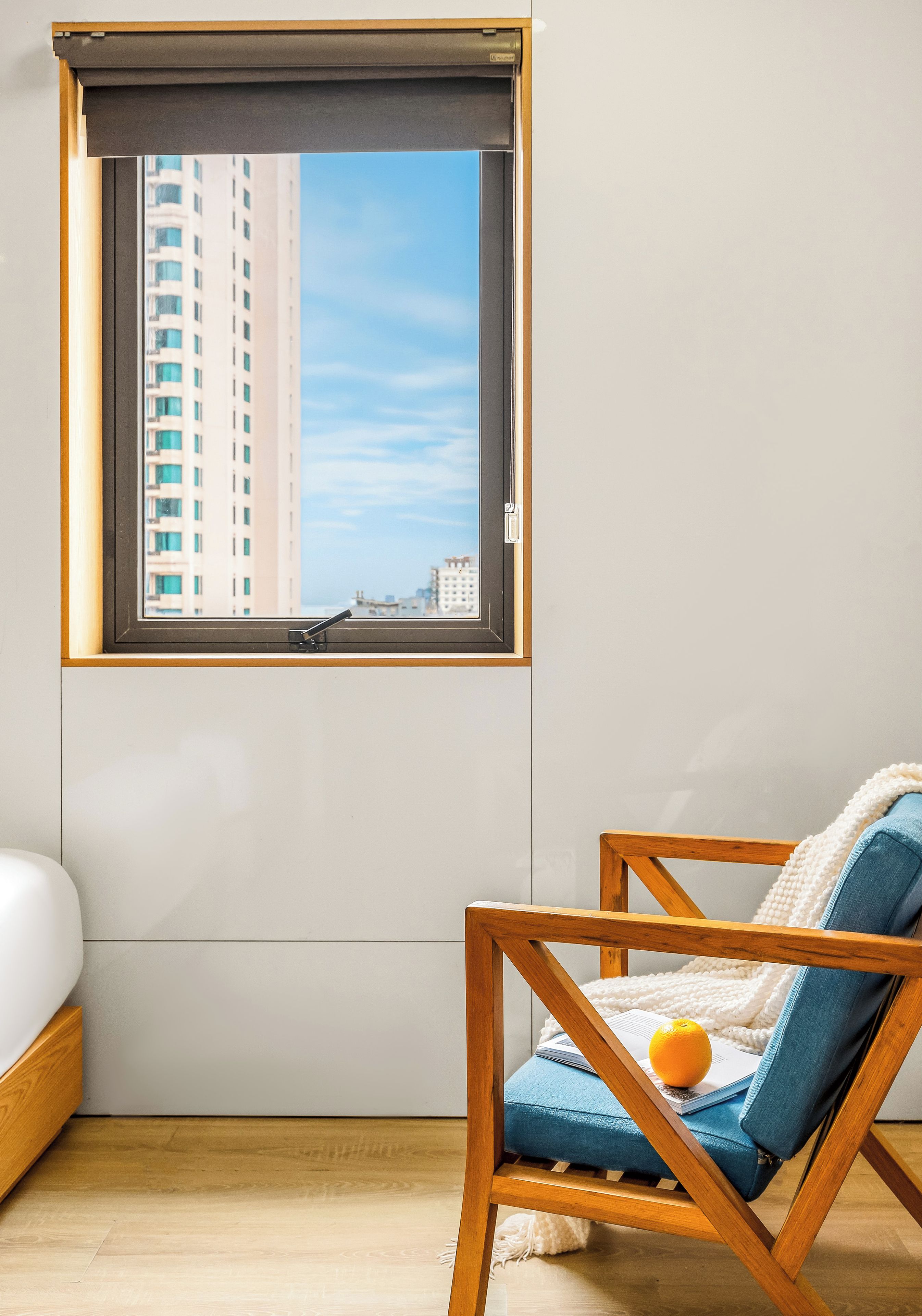 Ein Zimmer mit Fensterblick auf eine Stadtlandschaft, ein Holzstuhl mit Kissen und ein weißer Nachttisch.