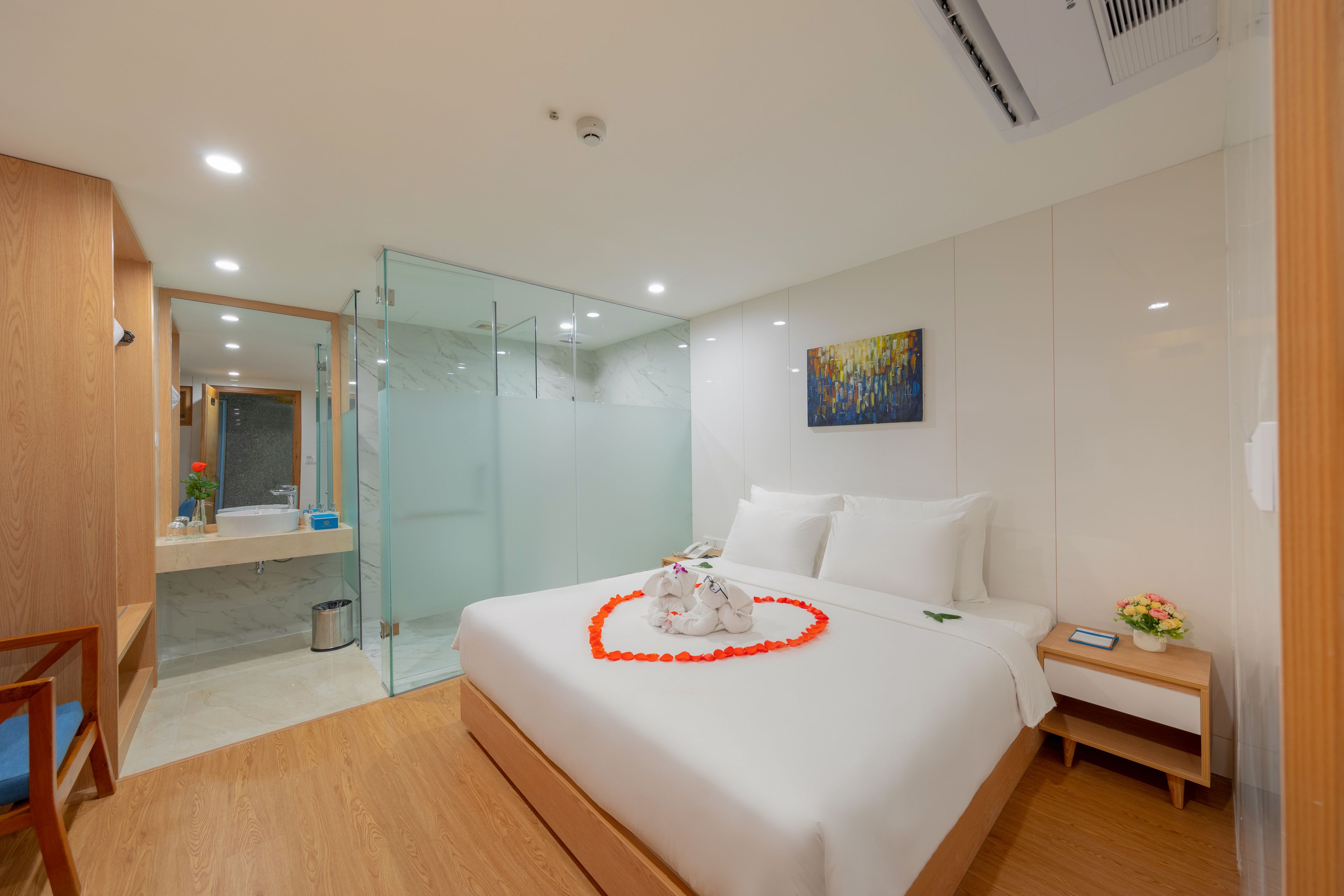 A modern hotel room with a large bed, a glass-enclosed shower, a wooden chair, and a small bedside table with a vase of flowers.