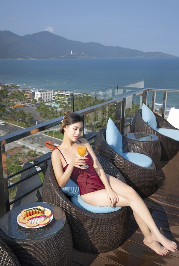 A woman relaxing on wicker lounge chairs with a glass of orange juice and a plate of fruit on a balcony overlooking a city and the sea. - KOVA Diamond Hotel (Da Nang)