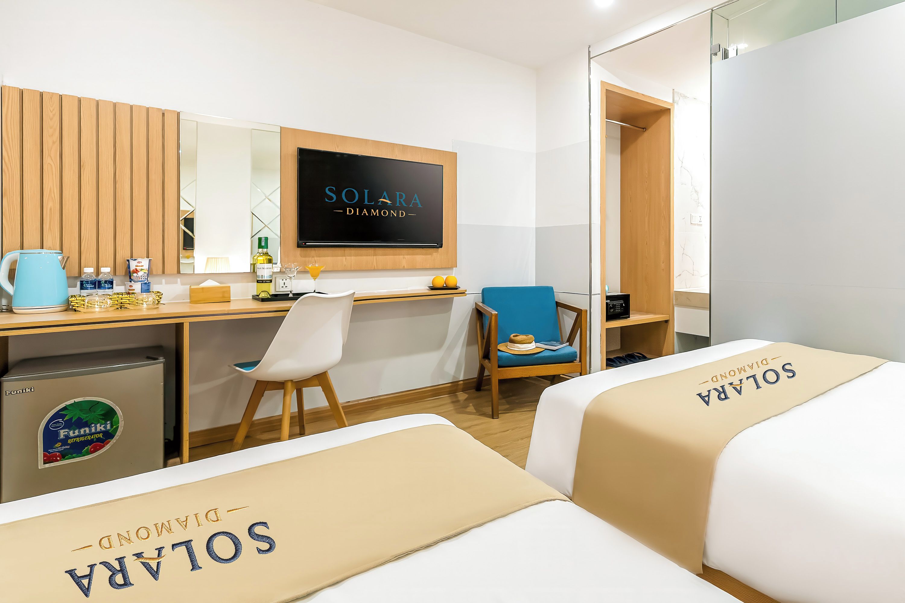 Habitación de hotel con cama, escritorio, silla, minibar y televisor mostrando 'SOLARA DIAMOND'.