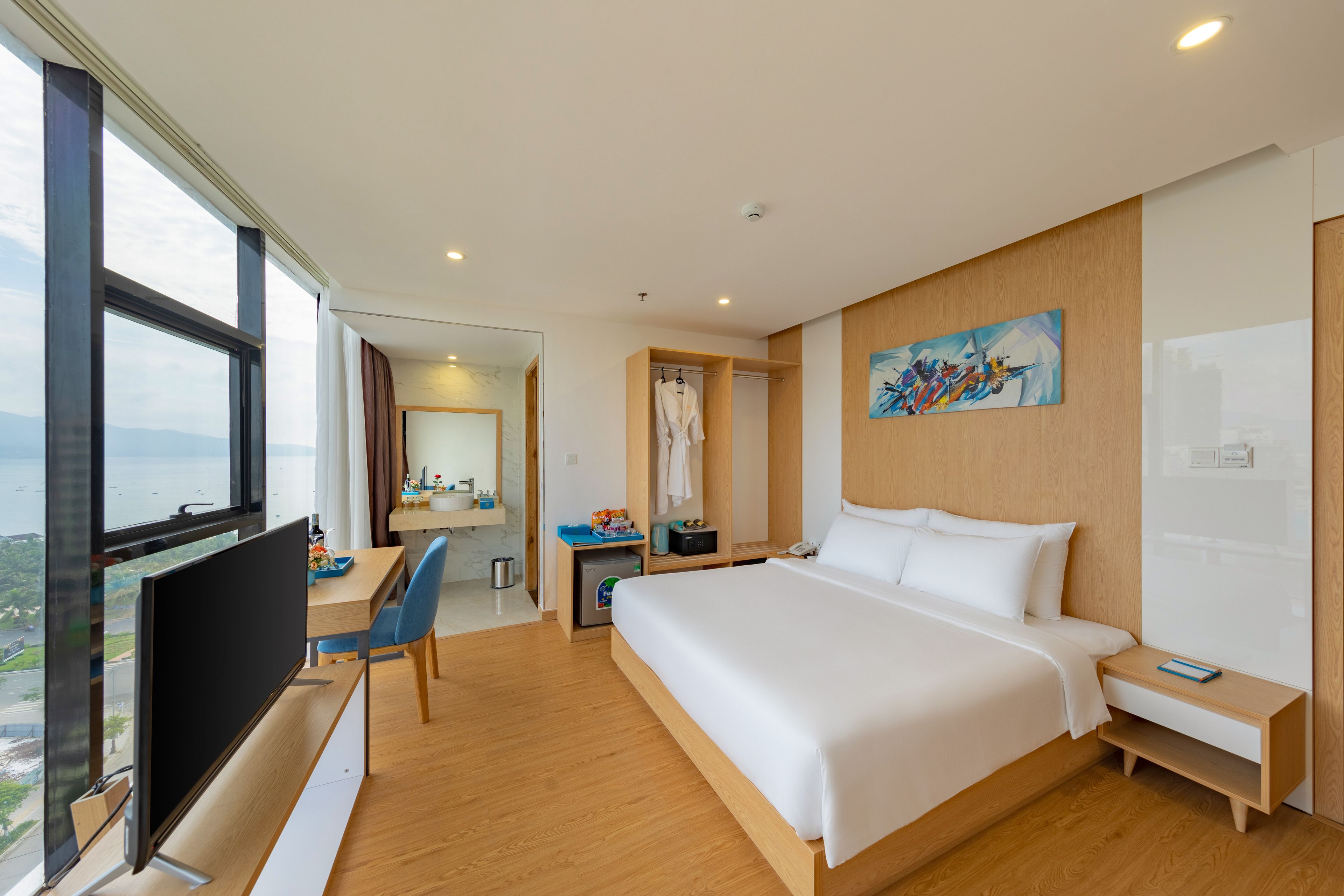 A modern hotel room with a large bed, a desk with a chair, a television, and a view of the sea.