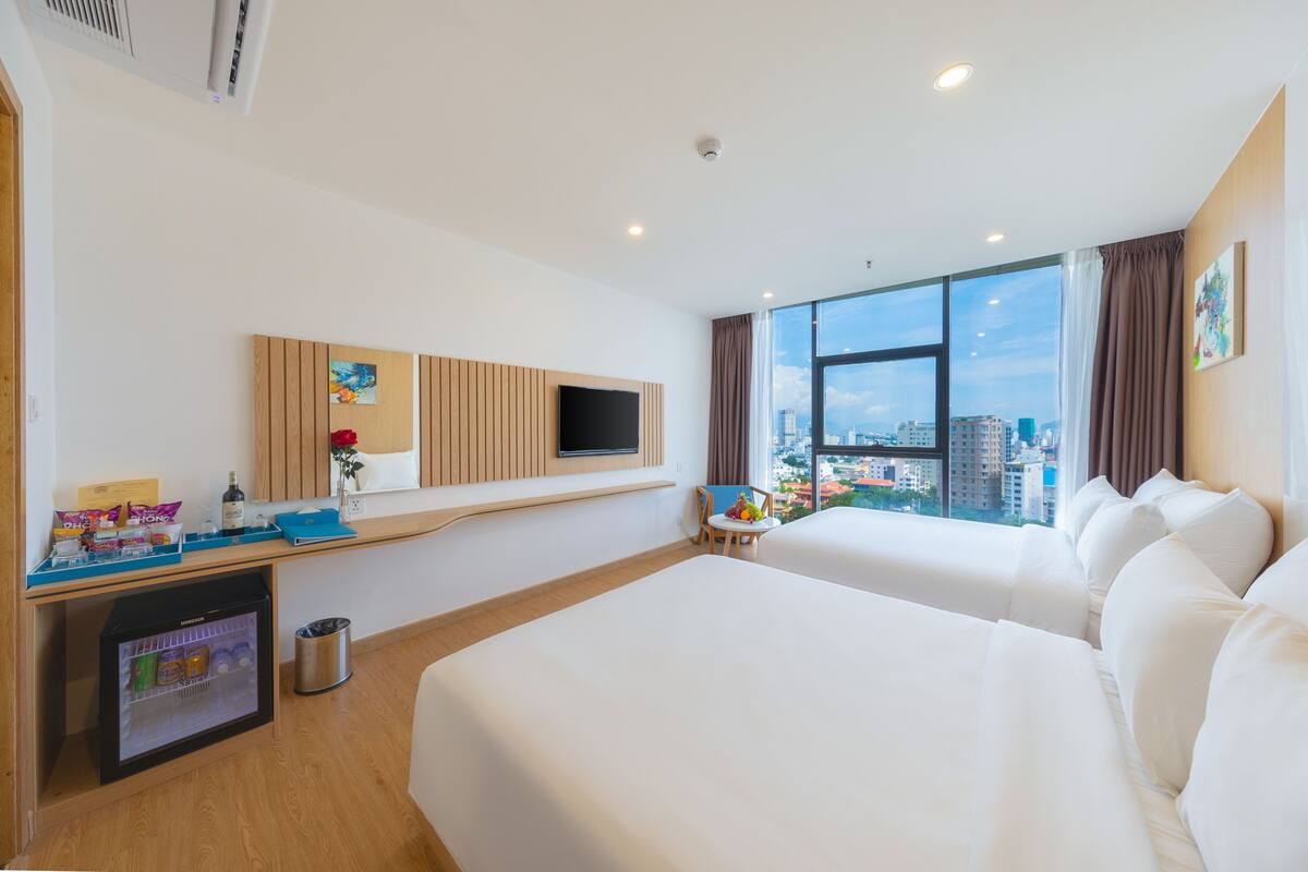 A hotel room with two beds, a TV, a mini-fridge, a small table with snacks, and a city view.