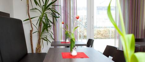 A modern dining area with a long table, black chairs, and a red placemat. There are two large potted plants and a vase with a single red tulip on the table.