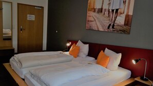 Una cama doble con ropa de cama blanca y almohadas naranjas, una mesita de noche con lámpara, y una puerta con un cartel.