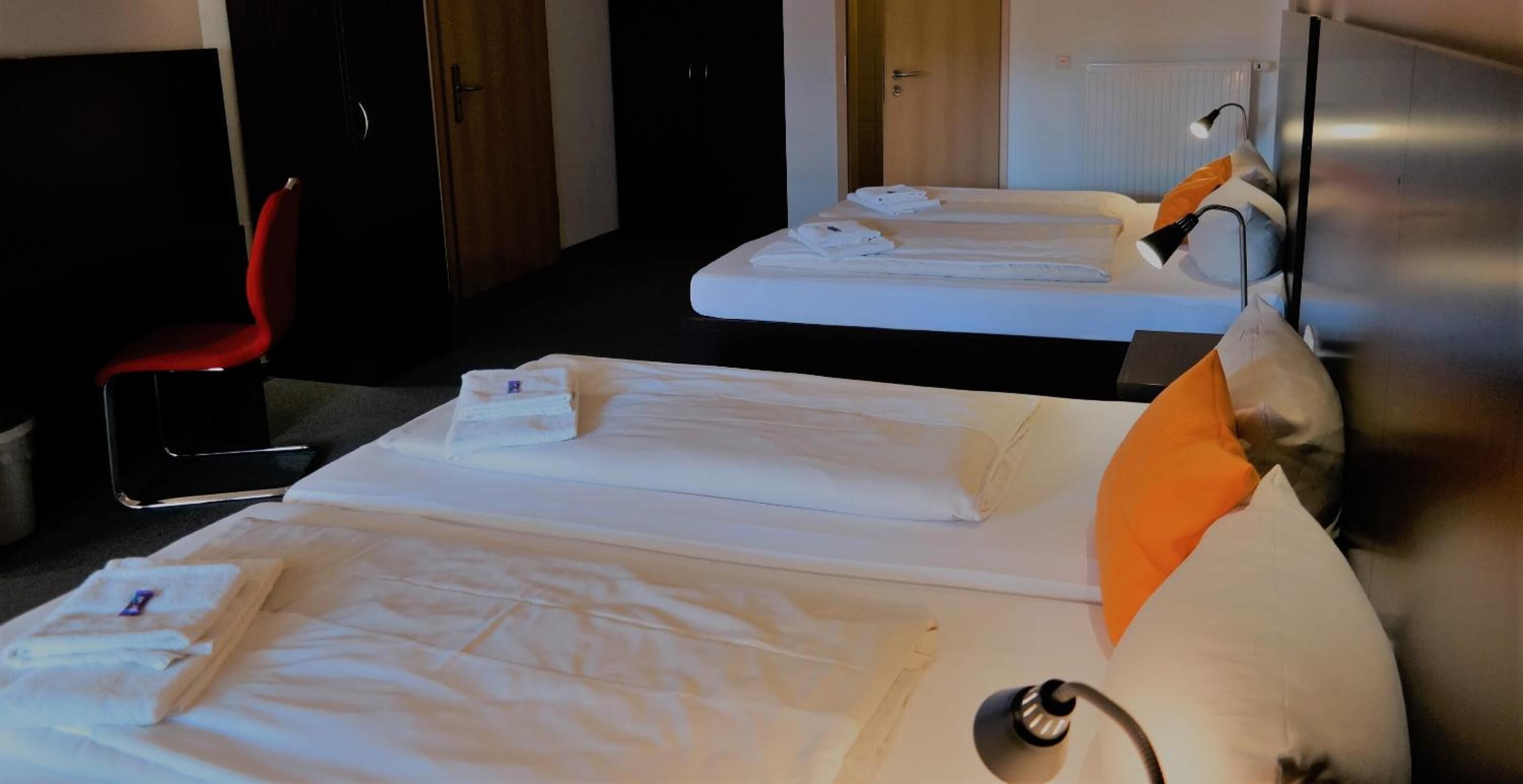 A hotel room with two beds, each with white linens and pillows, a red chair, and a bedside table with a lamp.