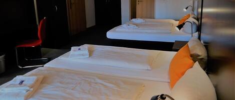 Habitación de hotel con dos camas, cada una con sábanas y almohadas blancas, una silla roja y una mesita de noche con lámpara.