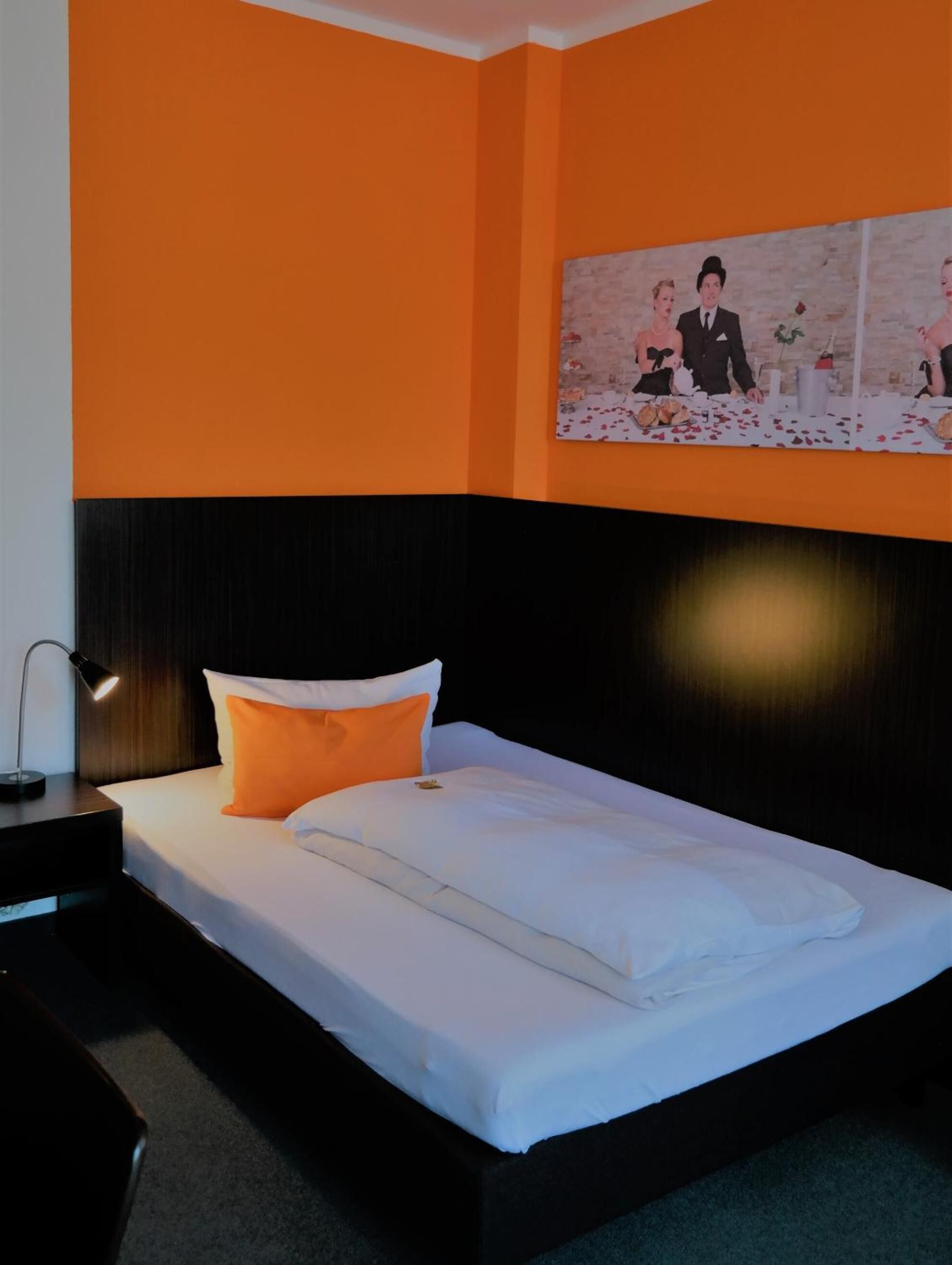A hotel room with an orange accent wall, a black headboard, and a bed with white bedding and an orange pillow.