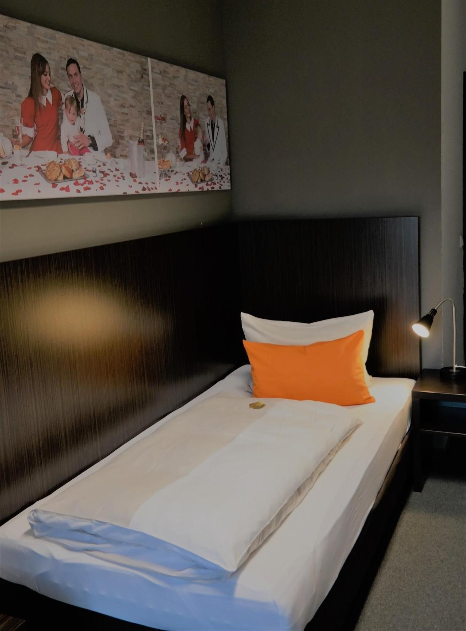 Un lit bien fait, avec des oreillers blancs et orange, une tête de lit sombre et une photo encadrée au-dessus.