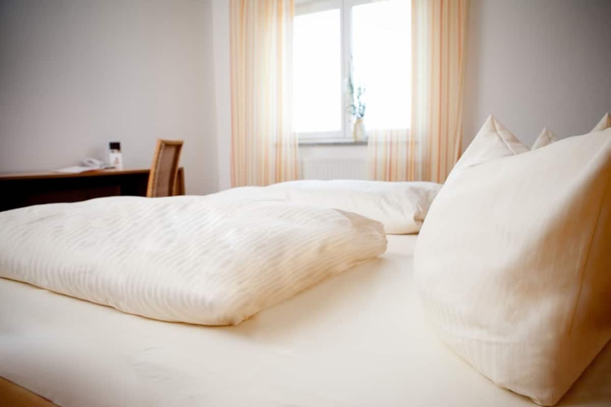 Una cama con ropa de cama y almohadas blancas, una mesita de noche de madera y una ventana con cortinas.