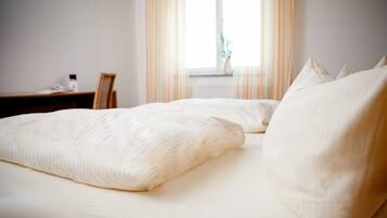 Una cama con ropa de cama y almohadas blancas, una mesita de noche de madera y una ventana con cortinas.