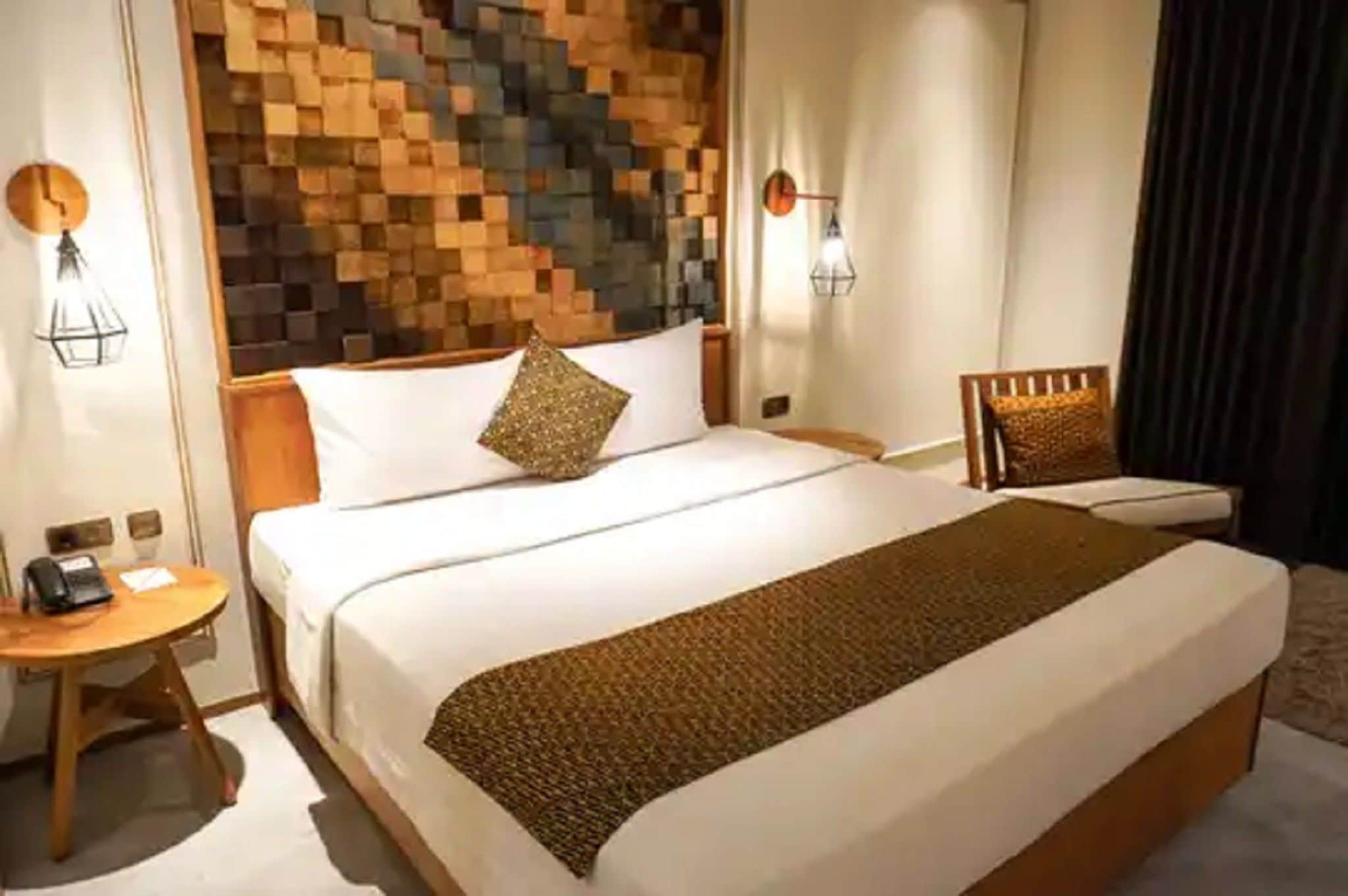A hotel room with a large bed, a bedside table, a chair, and a wall with a geometric pattern.