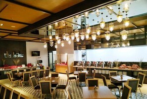 A restaurant with multiple dining tables and chairs, hanging pendant lights, and a kitchen area visible through a glass partition. - de Java Hotel Bandung (Bandung)