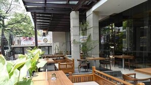 An outdoor cafe area with wooden tables and benches, surrounded by glass walls and potted plants. - de Java Hotel Bandung (Bandung)